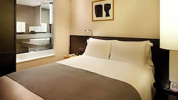 Habitación de hotel con una cama grande, una mesita de noche con lámpara, un tocador, un espejo y una obra de arte enmarcada en la pared.