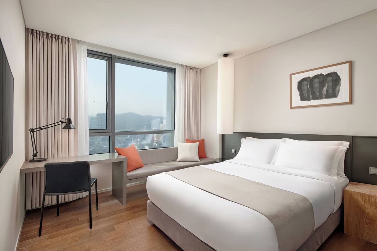 Habitación de hotel con cama, escritorio, silla, ventana con cortinas y un cuadro en la pared.