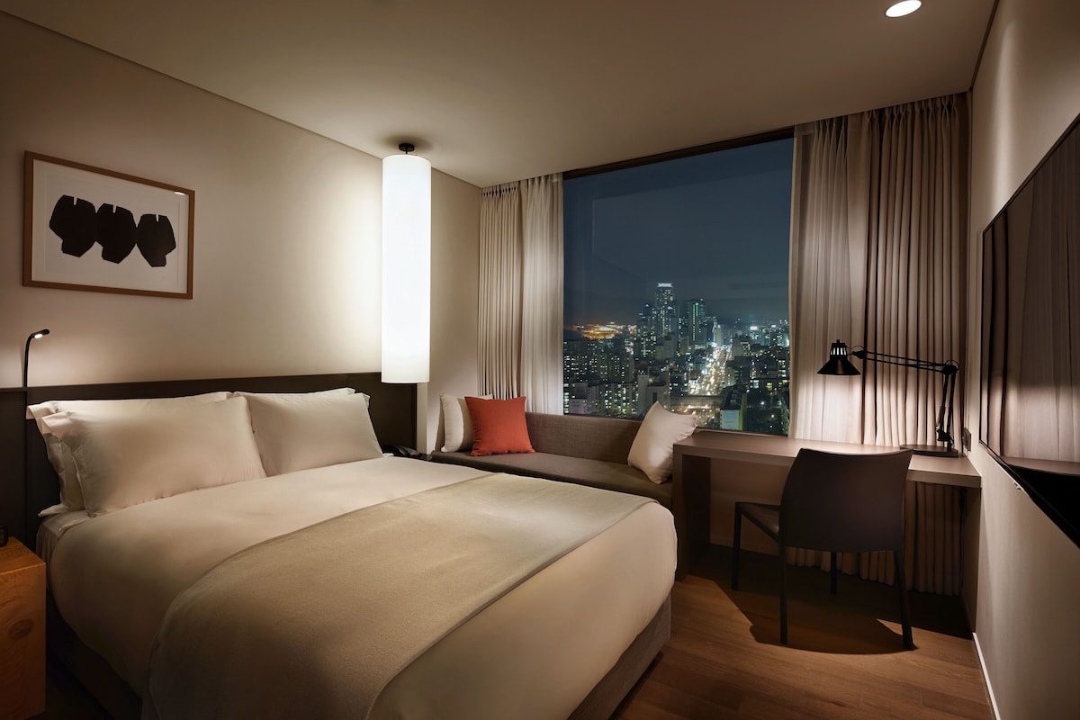 A hotel room with a large bed, a desk, and a view of a cityscape at night.