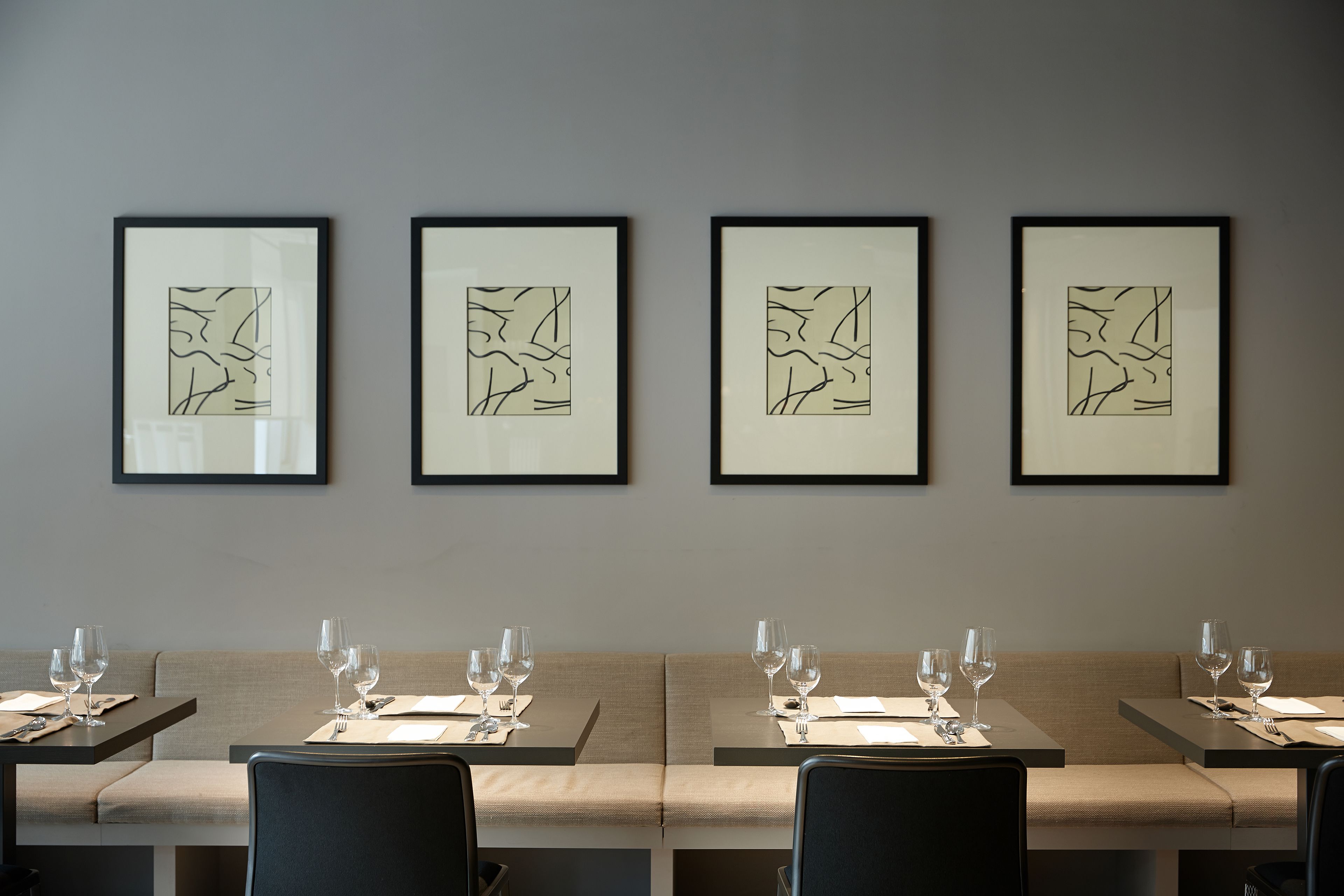 Um restaurante com quatro obras de arte abstratas emolduradas na parede, mesas preparadas com copos de vinho e talheres, e uma paleta de cores neutras.