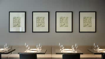 Um restaurante com quatro obras de arte abstratas emolduradas na parede, mesas preparadas com copos de vinho e talheres, e uma paleta de cores neutras.