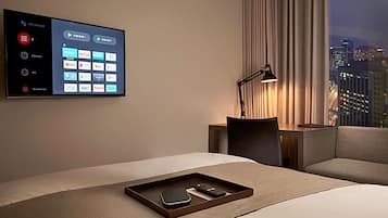 Habitación de hotel con una amplia pantalla digital que muestra diferentes servicios de streaming, una mesita de noche con lámpara y vistas a la ciudad por la ventana.