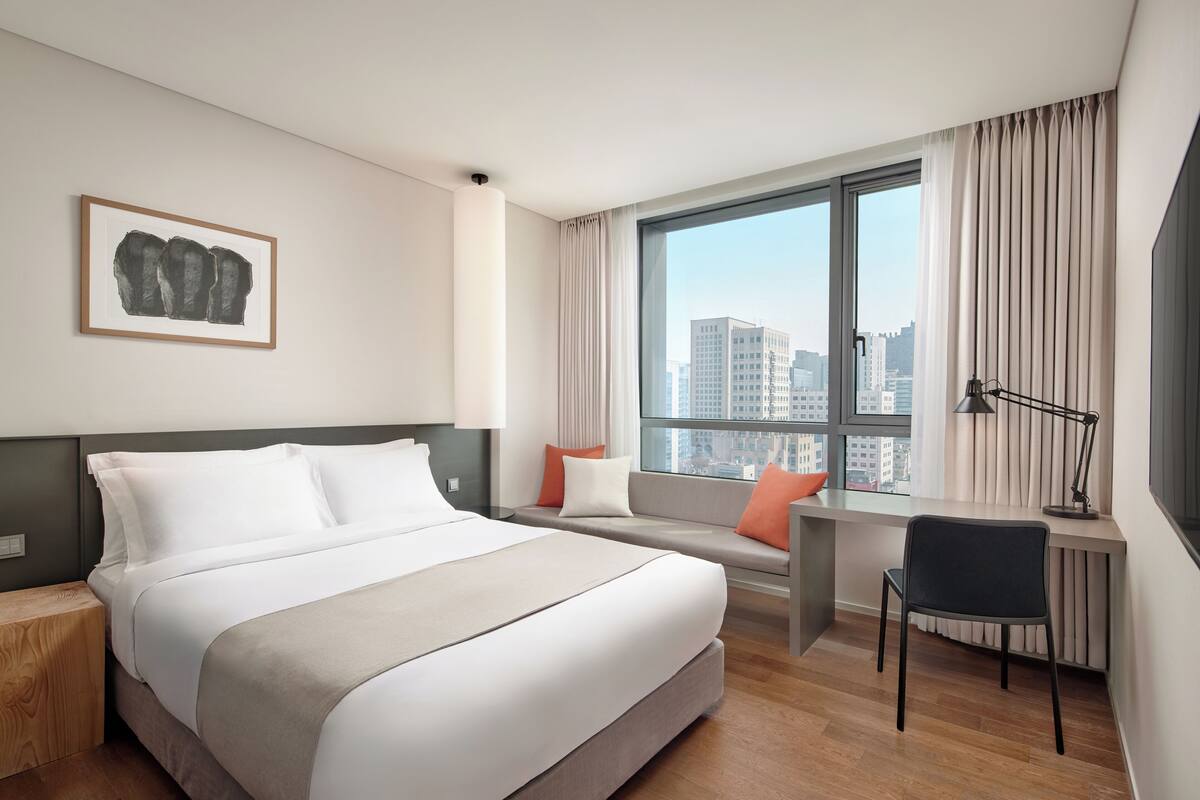 Una habitación de hotel con cama, escritorio, silla, ventana con cortinas y vista a la ciudad.
