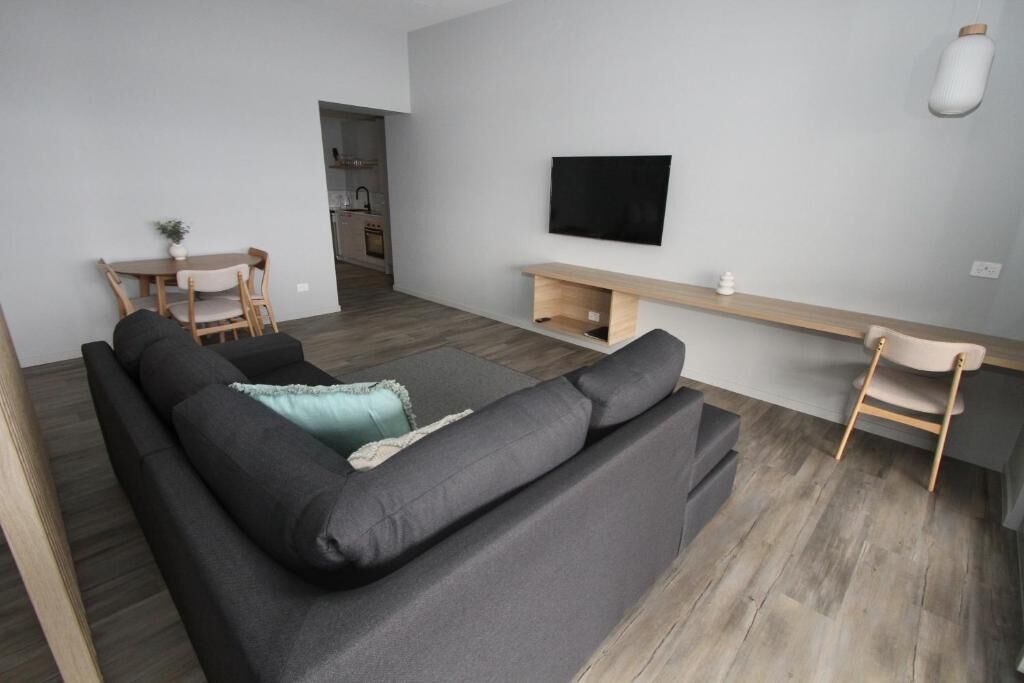 Una sala moderna con un sofá negro, un televisor montado en la pared y una mesa de comedor de madera con sillas.