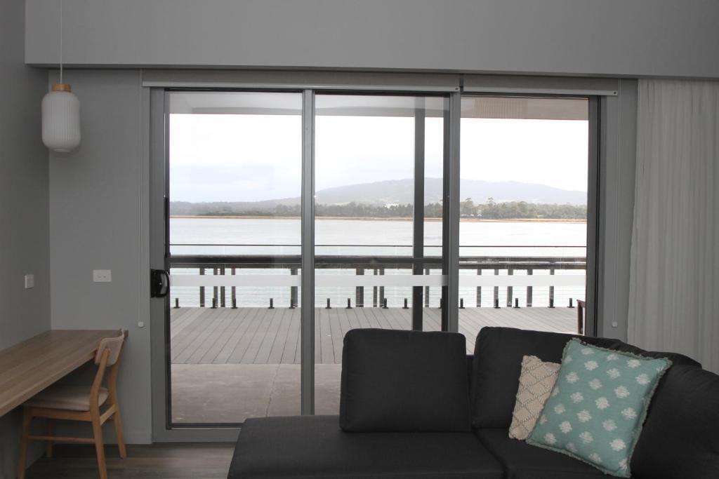 A modern living room with a black sofa, a wooden table, and a view of a body of water through large sliding glass doors.