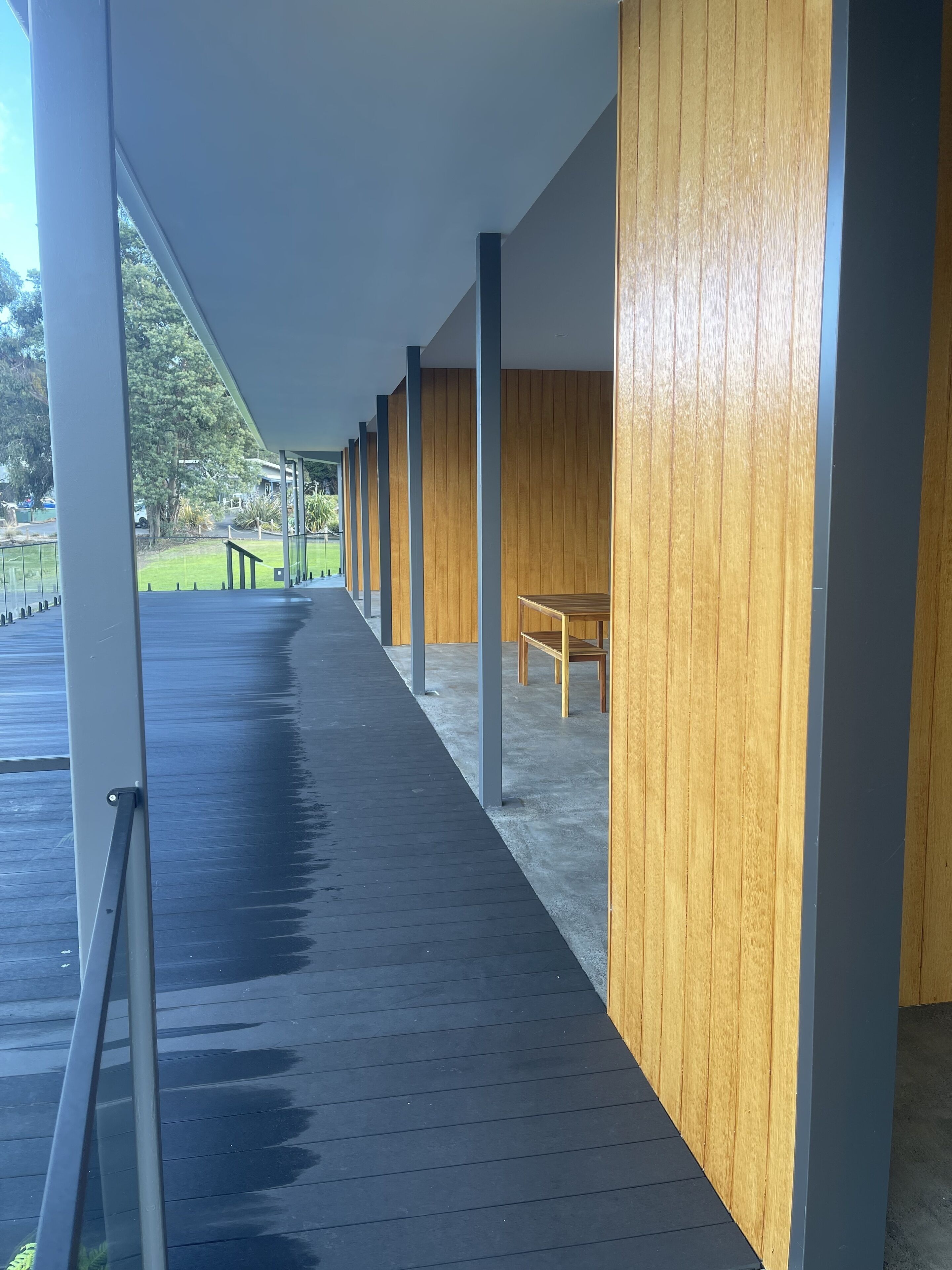A covered walkway with wooden paneling and benches, leading to a grassy area with a fence and trees.