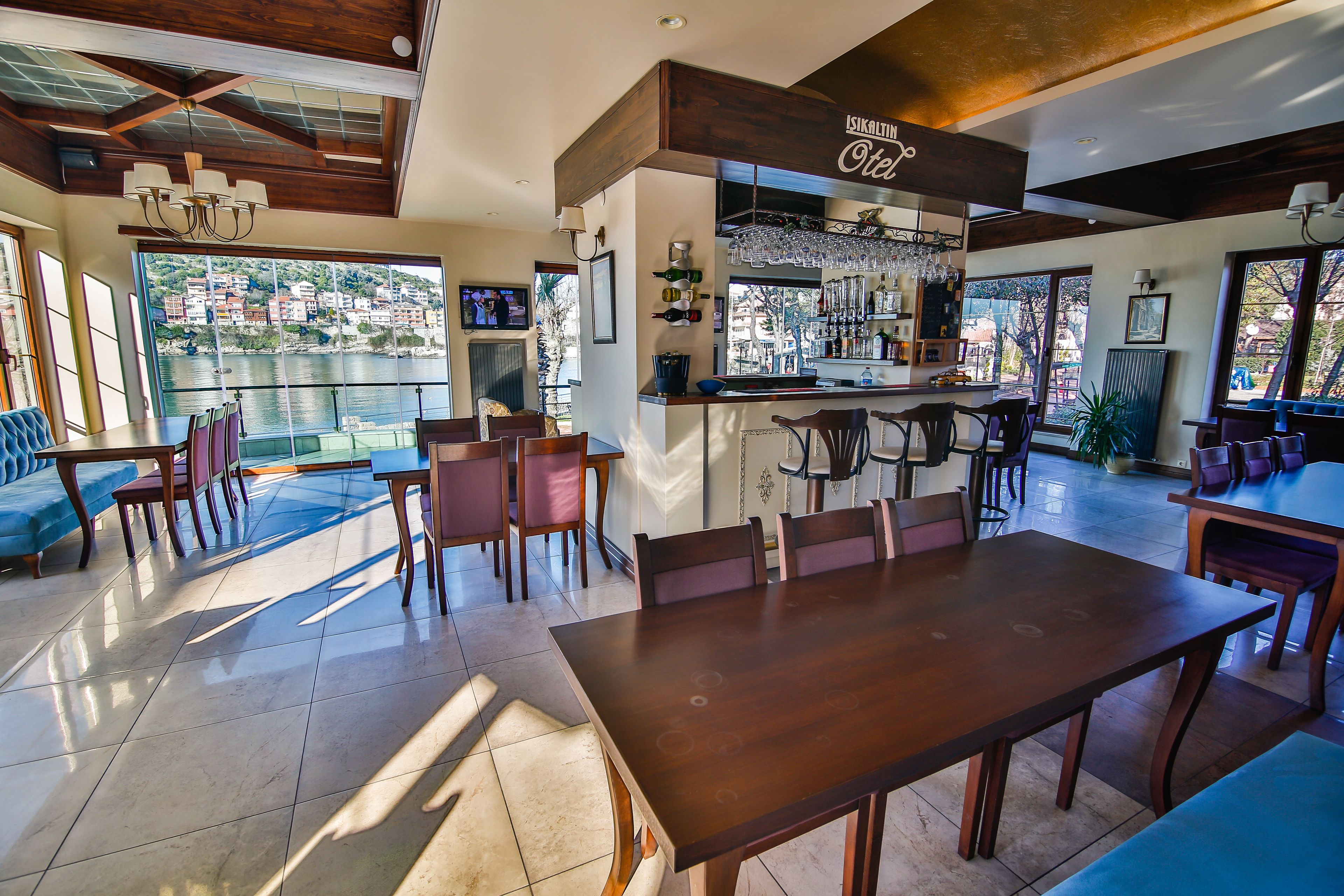 A spacious dining area with a large wooden table and chairs, a bar with wine glasses and bottles, and a view of the water and buildings outside.