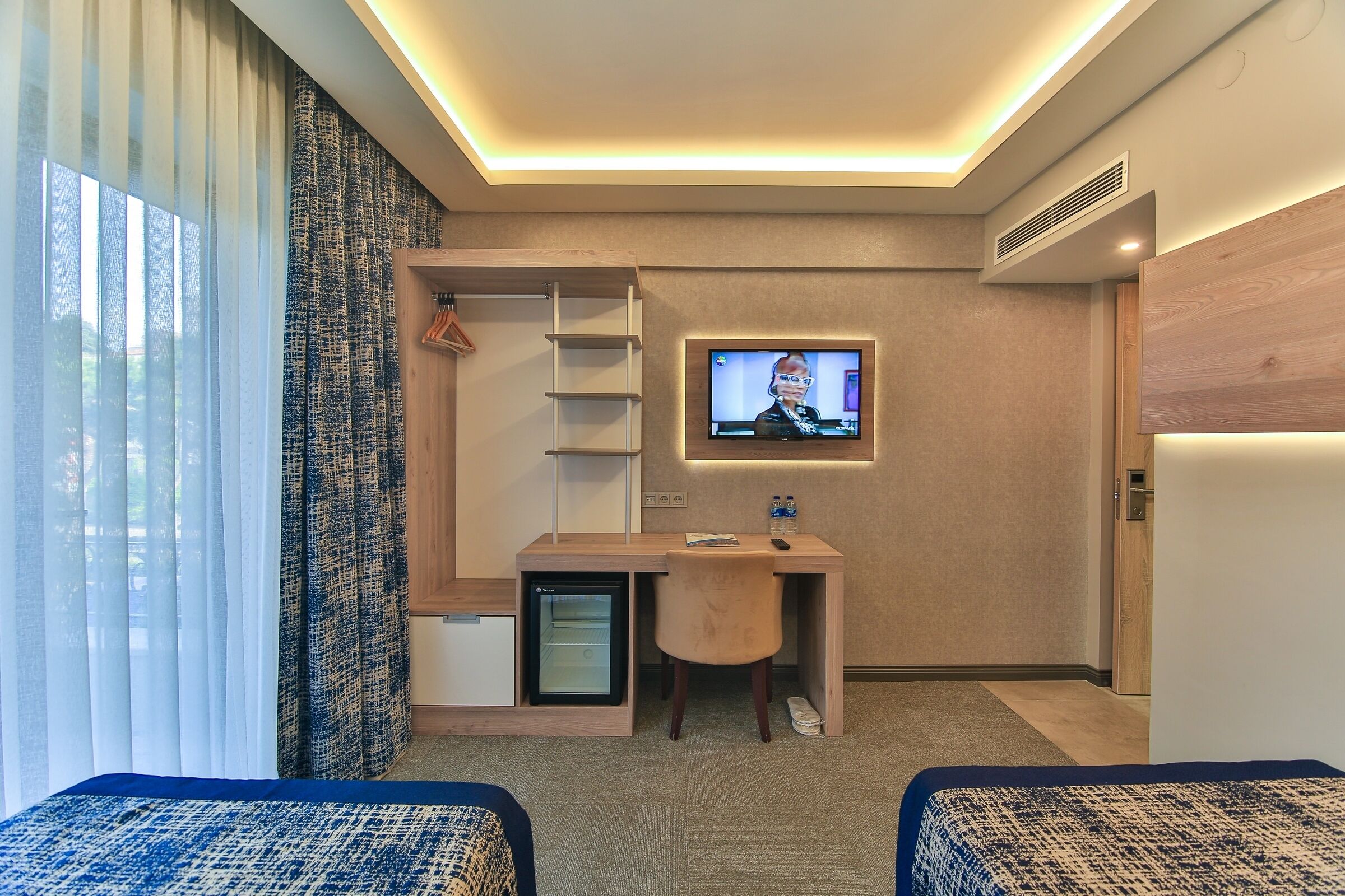 A hotel room with a bed, a desk with a TV, a mini-fridge, and a wardrobe.