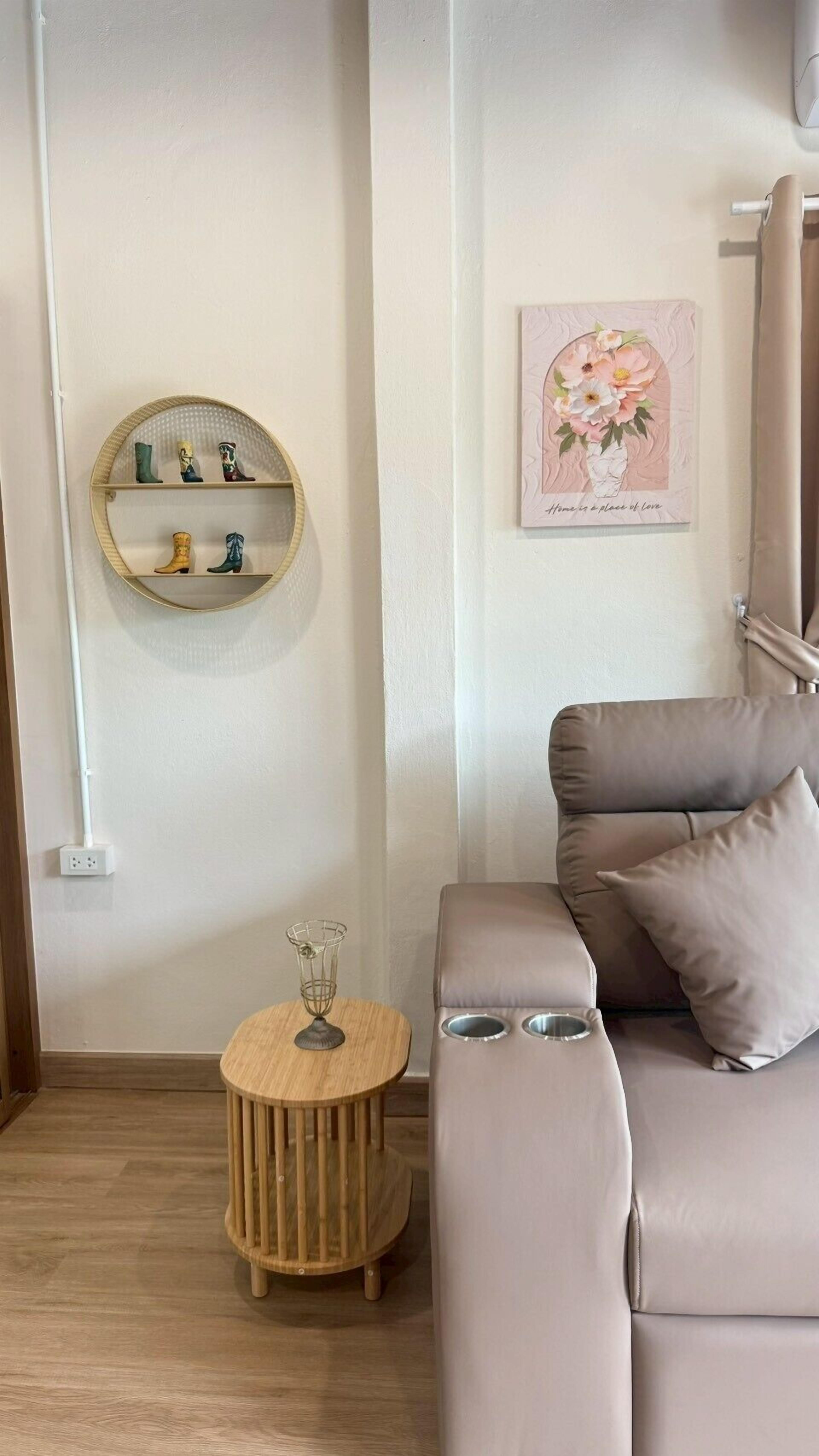 Een moderne woonkamer met een beige bank, een rond houten bijzettafeltje, een wandplank met decoratieve objecten, en een bloemrijk kunstwerk in een lijst.