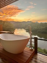 Un baño moderno con una bañera independiente, suelo de madera y vistas al río y las montañas durante el atardecer.