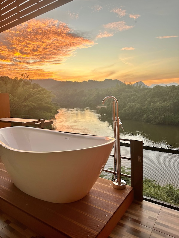 Un baño moderno con una bañera independiente, suelo de madera y vistas al río y las montañas durante el atardecer.