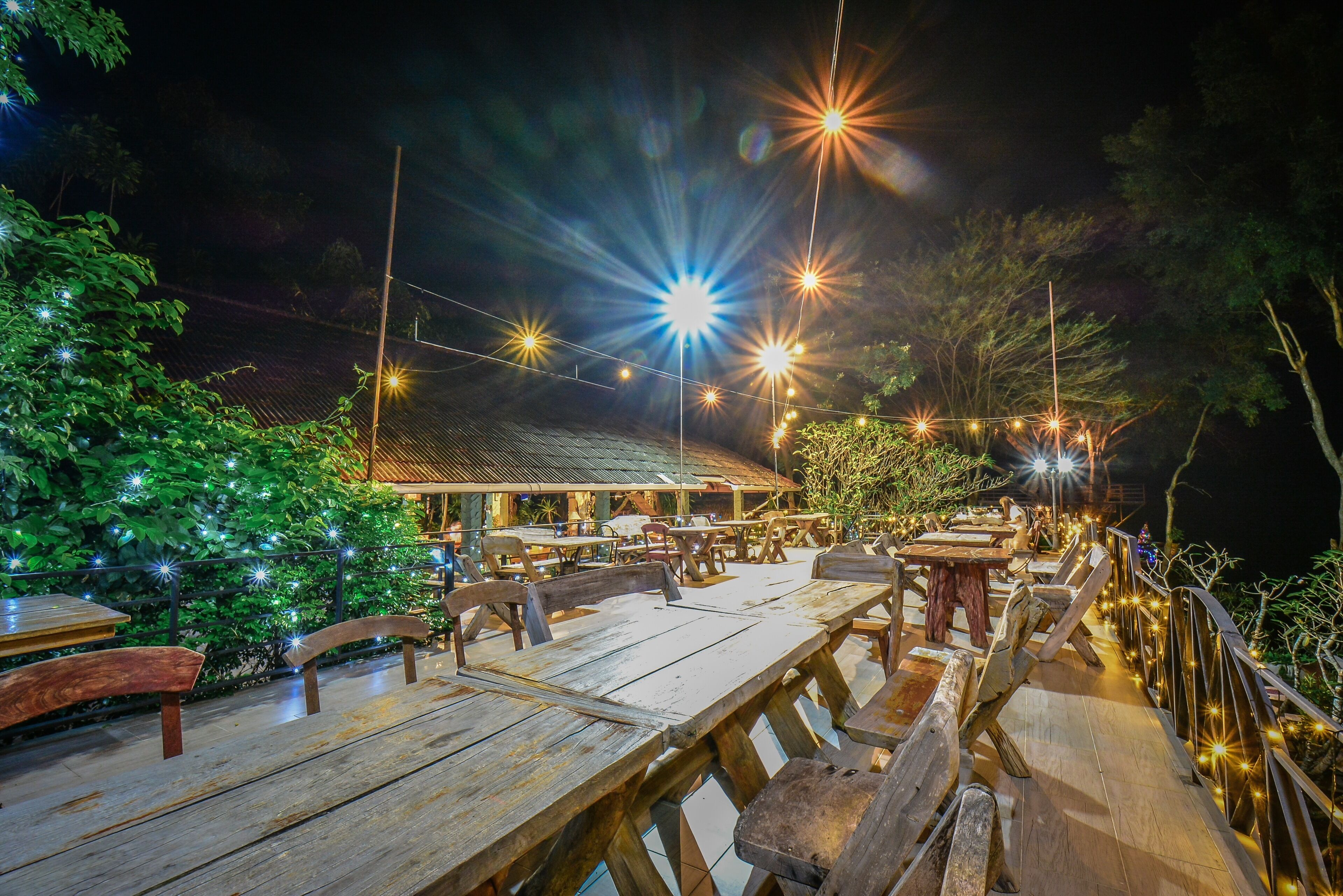 Een terras met houten tafels en banken, sfeerverlichting en een uitzicht op een gebouw op de achtergrond.