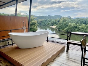 Un baño moderno con una bañera independiente, piso de madera y vistas a un río y colinas boscosas.