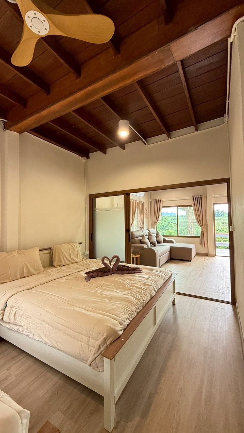 Ein Schlafzimmer mit einem großen Bett, Holzbalkendecke, Blick ins Freie durch eine Schiebetür und einem Deckenventilator.