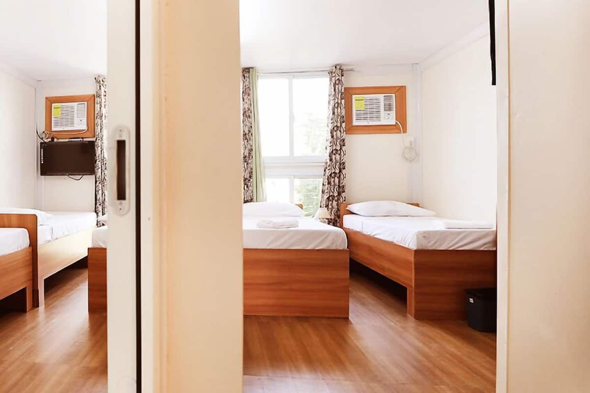 A hotel room with two wooden beds, a window with curtains, a wall-mounted air conditioner, and a small trash bin.