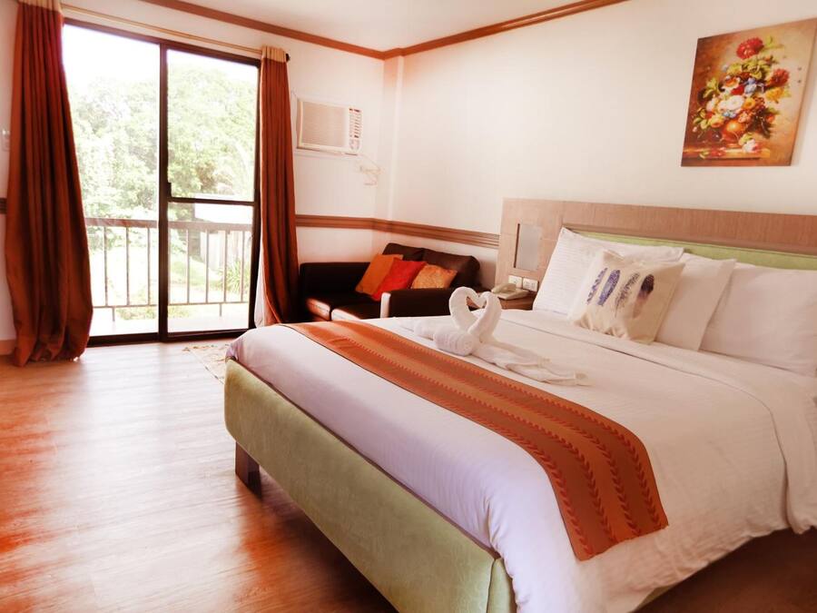 A hotel room with a large bed, a sofa, a balcony with curtains, and a painting on the wall.