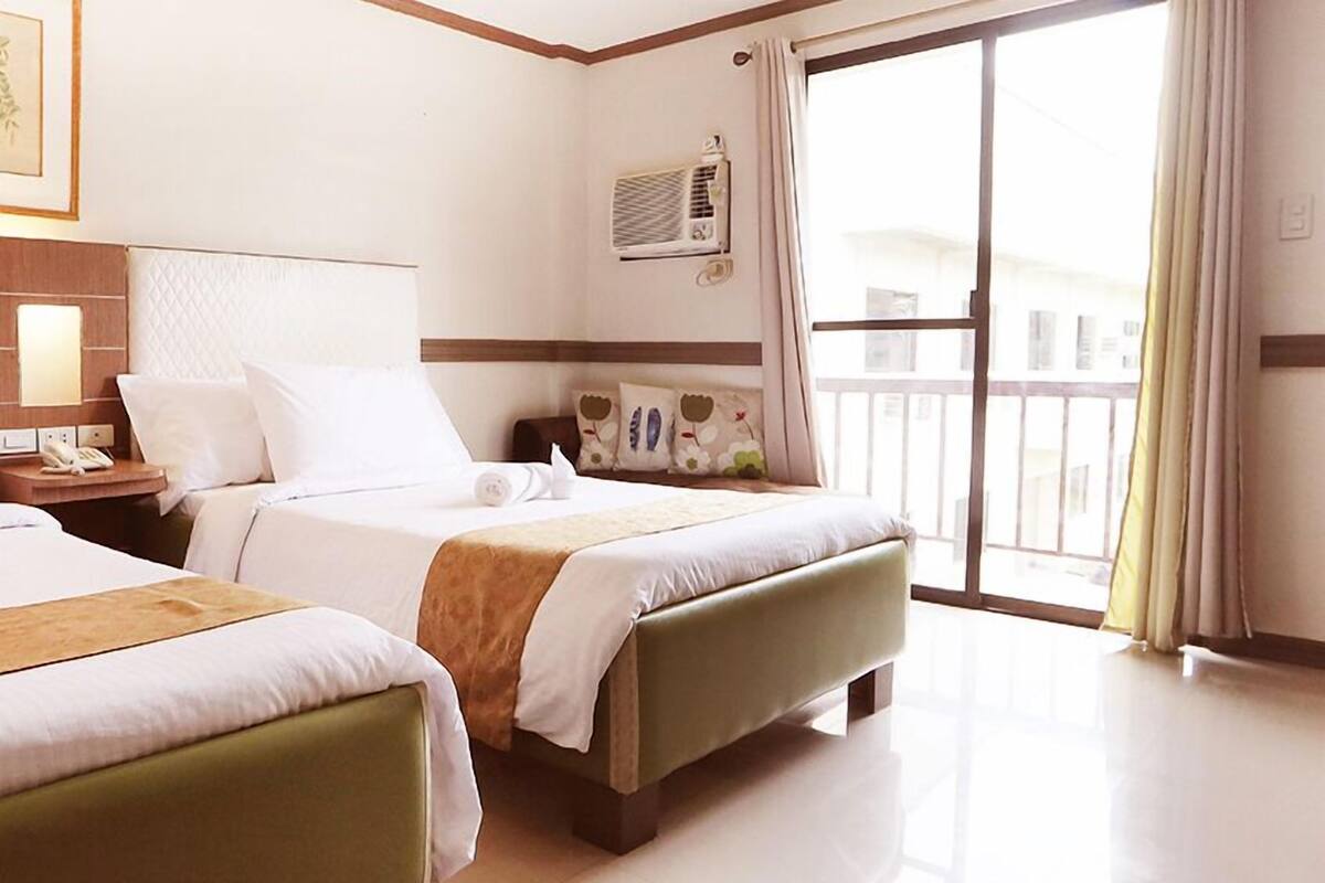 A hotel room with two beds, a bedside table with a lamp, a window with curtains, and a wall-mounted air conditioning unit.