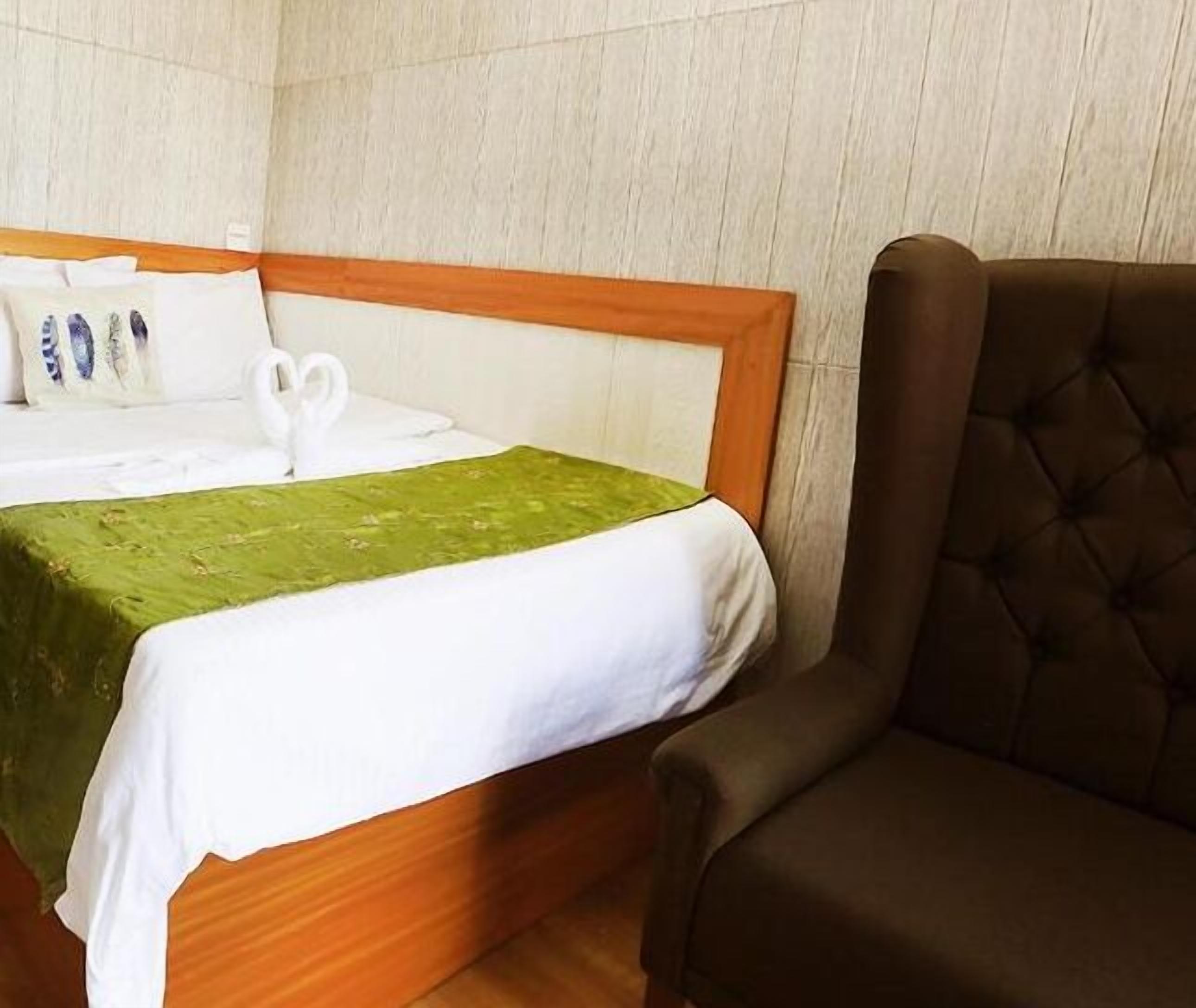 Una habitación de hotel con cama, cubrecama verde, almohadas blancas y un sillón marrón.
