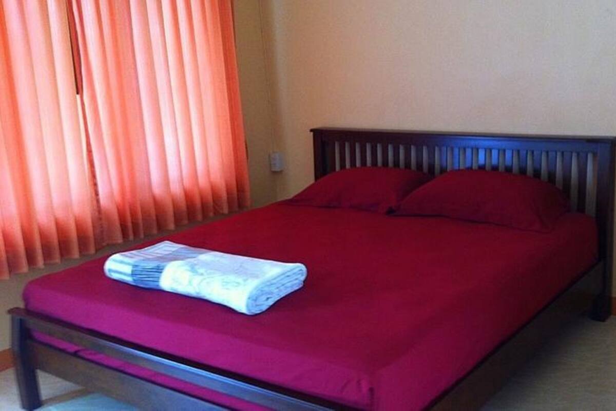 Een opgemaakt bed met rode beddengoed en kussens, een houten hoofdbord en een raam met oranje gordijnen.