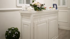 Un espace d’accueil propre et lumineux, aux murs blancs, où est affiché un panneau indiquant « Charme & Chic, chambre d’hôtes de luxe ». On y trouve un comptoir d’accueil orné d’un vase de fleurs, d’une plante en pot et d’un paillasson portant le même nom.