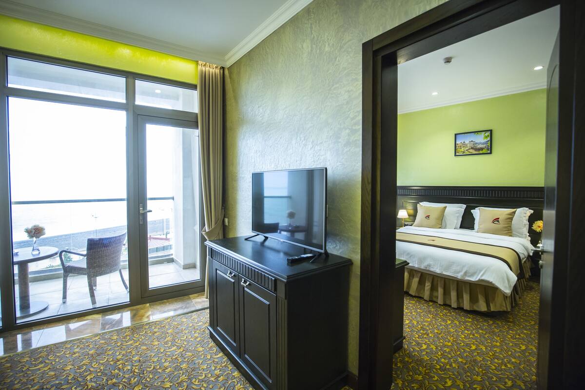 A hotel room with a large window, a television on a dresser, a bed with a headboard, and a painting on the wall.