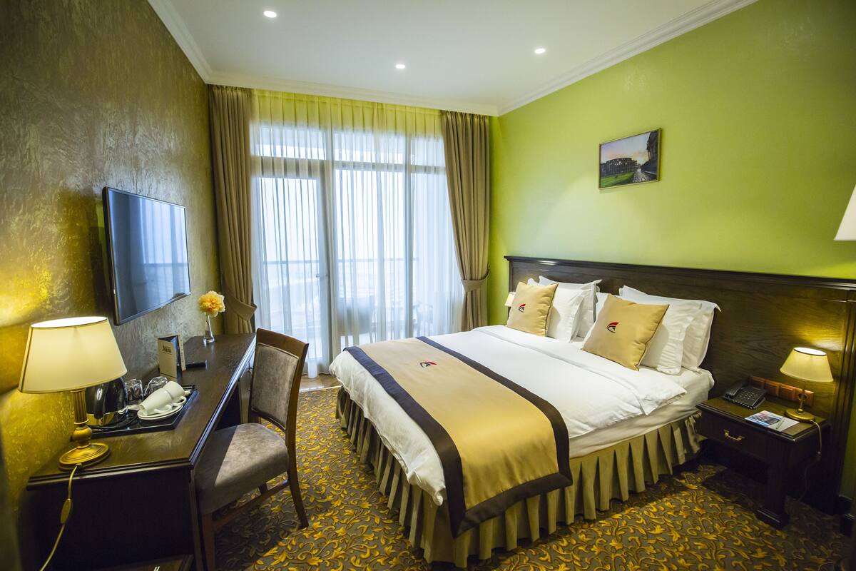 A hotel room with a large bed, a desk with a telephone, a chair, a television, and a nightstand with a lamp.