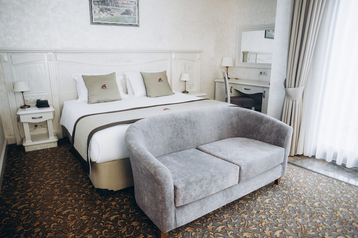 A hotel room with a bed, a grey sofa, a nightstand, a lamp, and a patterned carpet.
