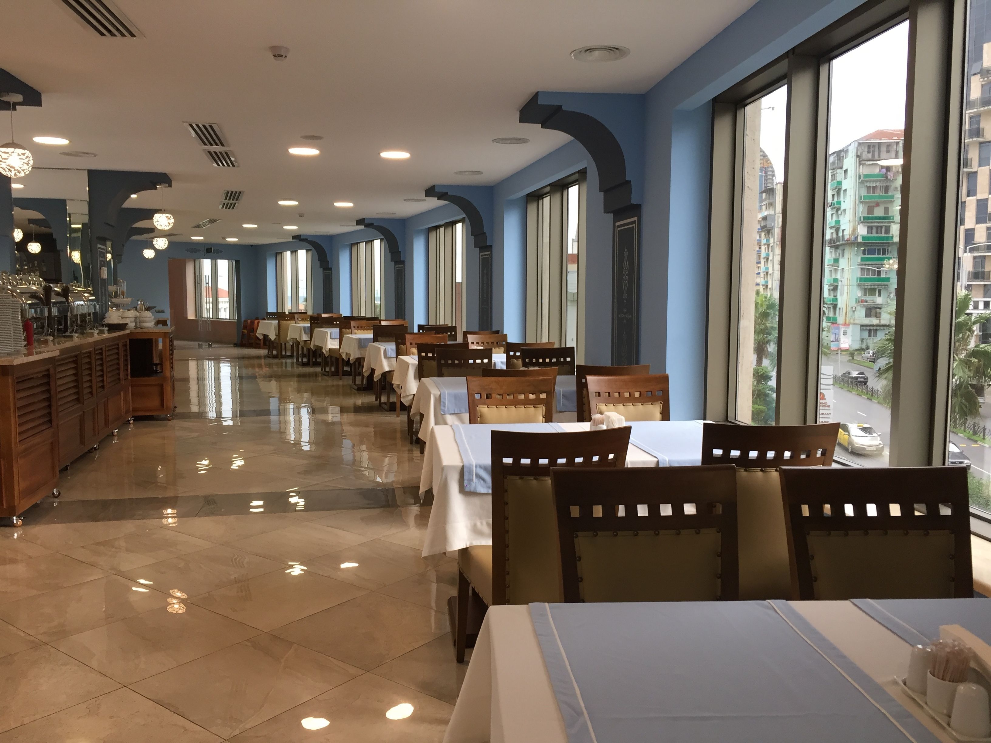 A restaurant with long tables and chairs, large windows, and a view of buildings outside.