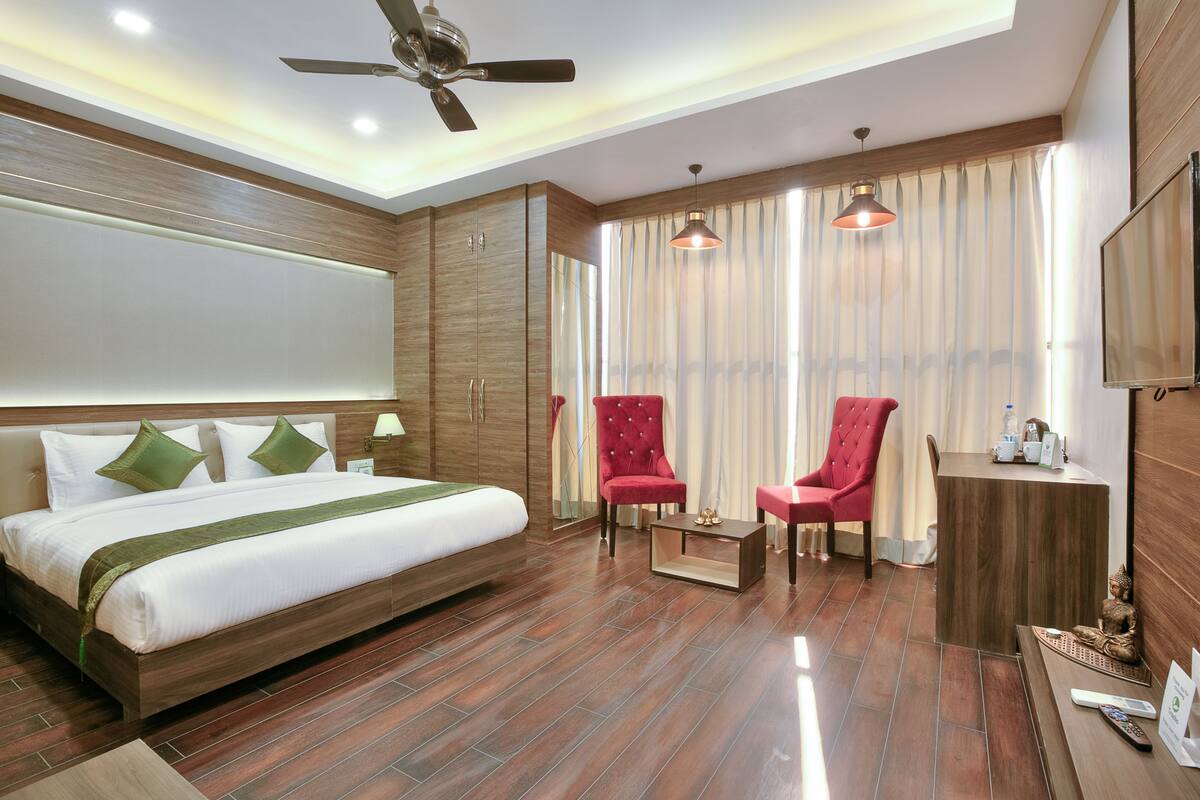 A hotel room with a large bed, two red armchairs, a small table, a ceiling fan, and a TV on the wall.