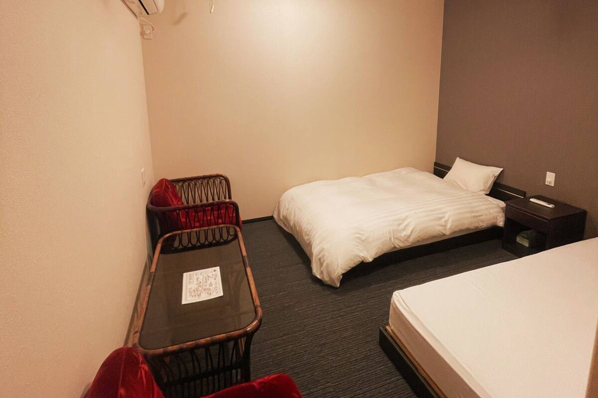Hotellihuone, jossa on yksi sänky, pieni pöytä, jossa on kortti, tuoli ja yöpöytä, jossa on kaukosäädin.