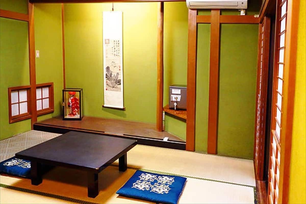 Tatami-mattoilla päällystetty huone, jossa on matala pöytä, kehystetty kuva ja pieni hylly, jonka päällä on jokin laite.