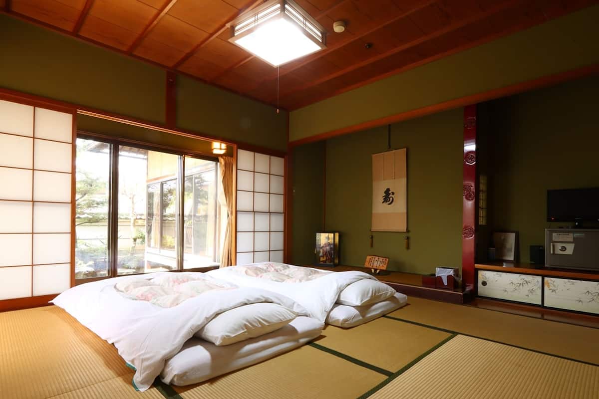 Perinteinen japanilaistyylinen huone, jossa on tatamimatto, suuri sänky valkoisilla vuodevaatteilla, liukuvat shoji-ovet ja televisio puukehikossa.