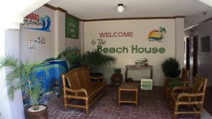 Un área de recepción con un letrero que dice 'Welcome to the Beach House', muebles de mimbre, plantas en macetas y un mural de una escena de playa.