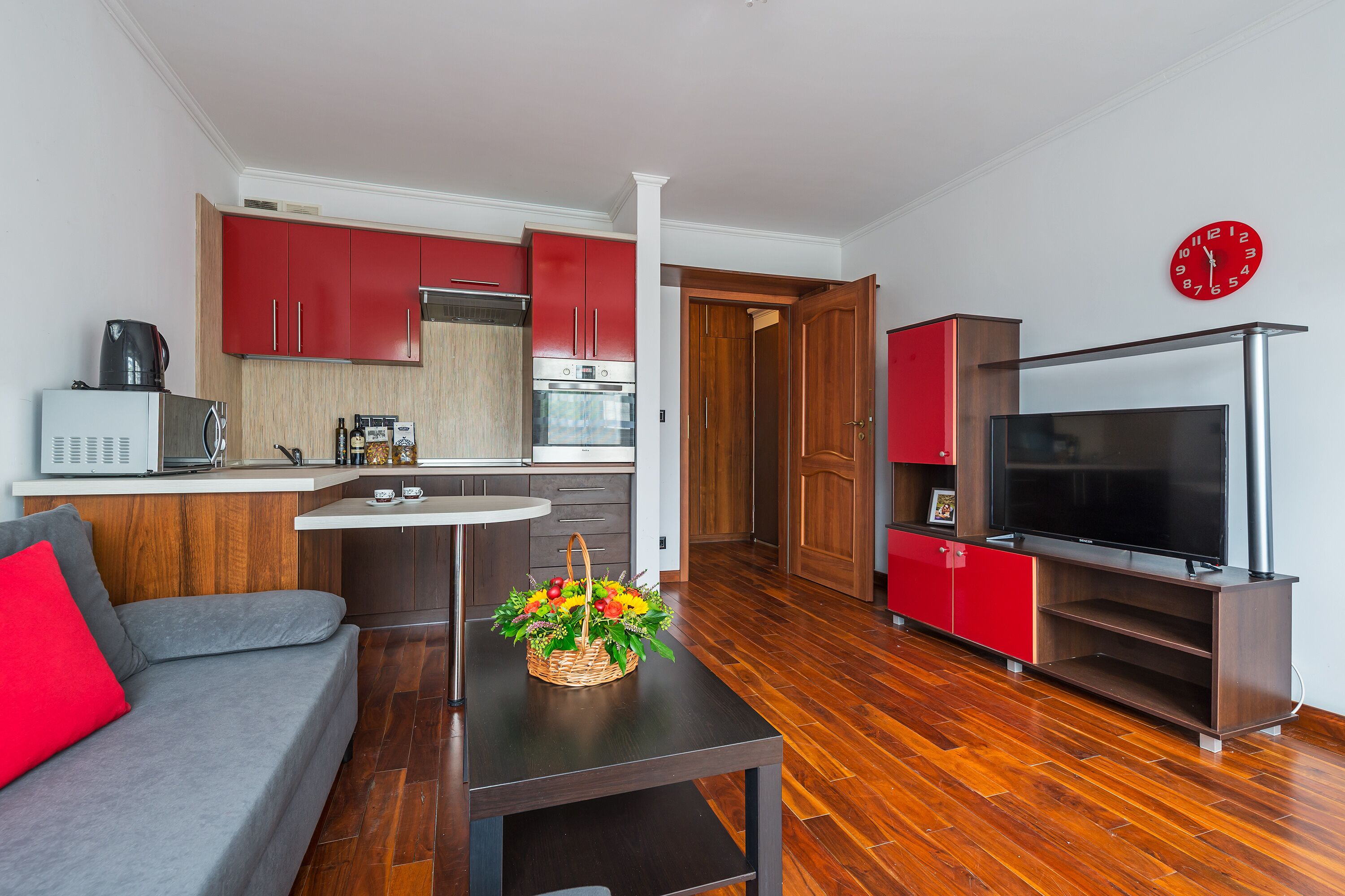 Uma sala de estar moderna com um sofá cinzento, uma mesa de centro de madeira com um arranjo de flores, um móvel para entretenimento vermelho e uma área de cozinha com armários vermelhos e um micro-ondas branco.