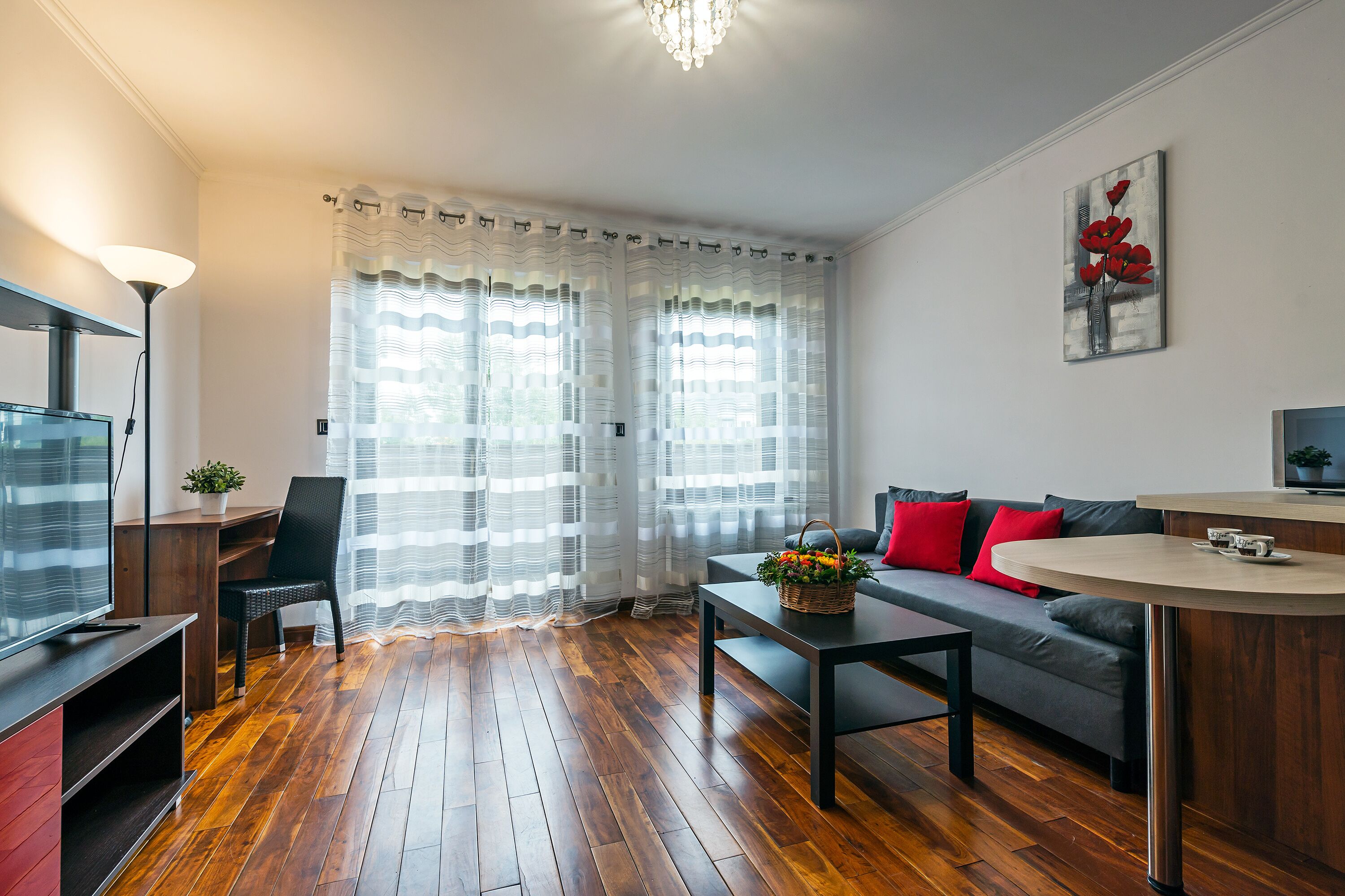 A modern living room with a wooden floor, a grey sofa with red cushions, a round coffee table, a TV on a stand, and a painting on the wall.