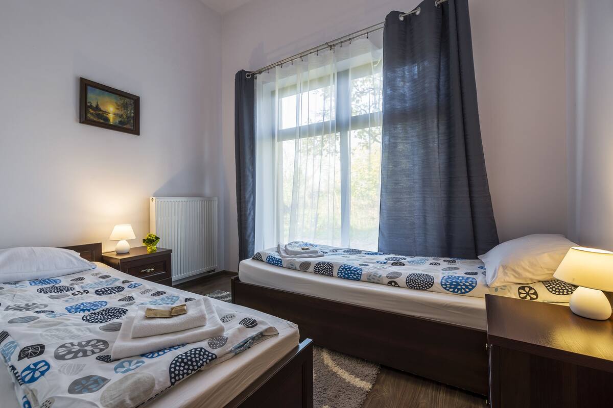 Une chambre d’hôtel avec deux lits, une fenêtre avec des rideaux, un radiateur, une table de chevet avec une lampe et un tableau encadré au mur.