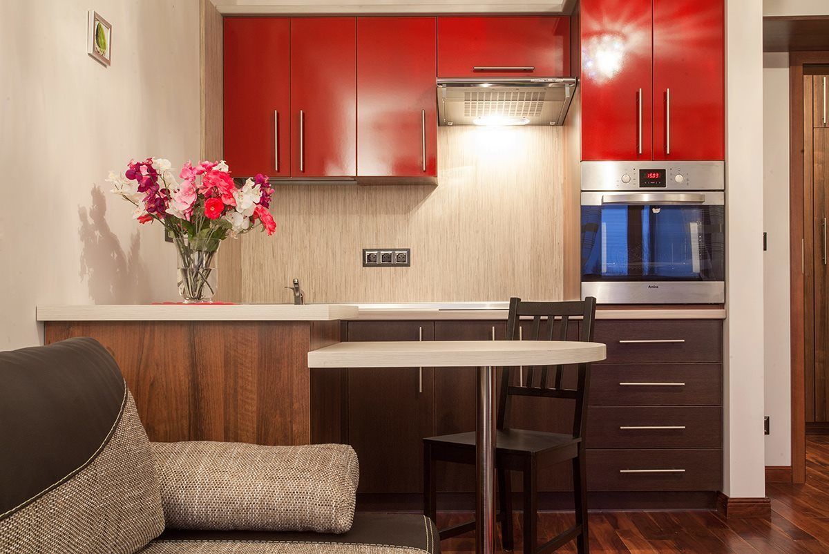 A compact kitchen with red cabinets, a stainless steel oven, and a dining area with a table and chairs.