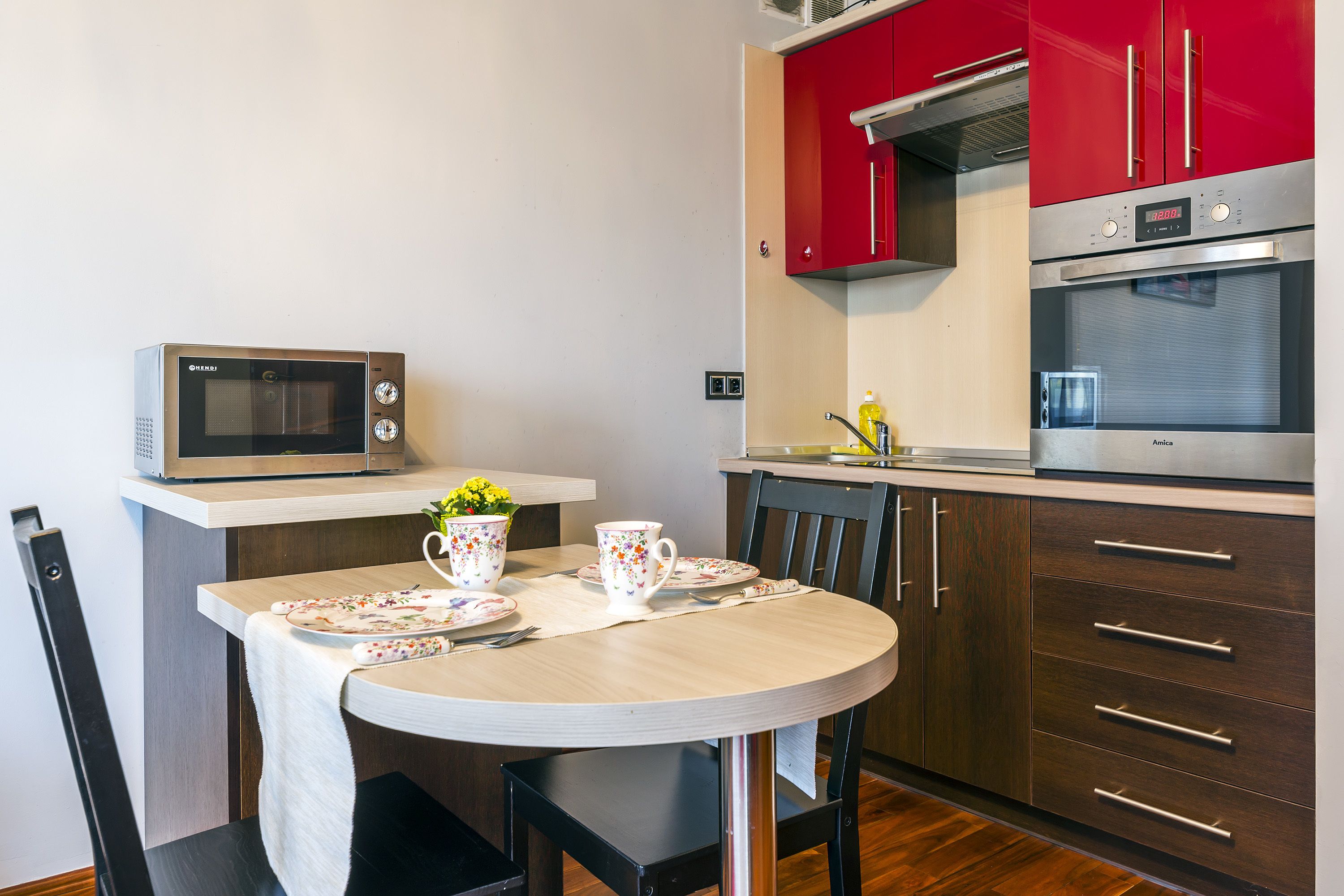 A kitchen with a microwave, oven, and red cabinets. There is a table with a floral tablecloth, a flower vase, and a coffee pot.