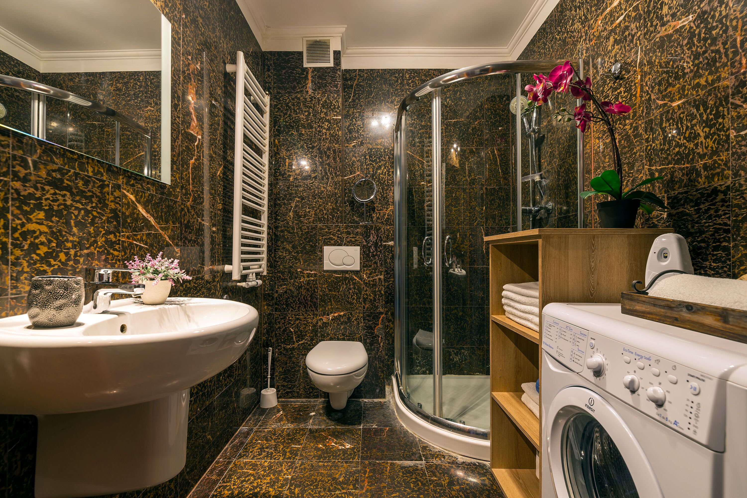 Um banheiro moderno com painel de fundo de mármore, chuveiro com box de vidro, máquina de lavar embutida e vaso sanitário.