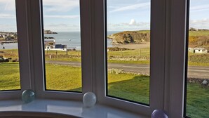 A room with a large window offering a view of a coastal landscape, including a beach and a body of water. - Castellor (Cemaes Bay)