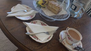 A dining table with a glass dome cake stand, a cup of tea, and a plate with a napkin and fork. - Castellor (Cemaes Bay)