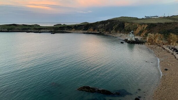A coastal landscape with calm waters, grassy hills, and a small white building on the right. - Castellor (Cemaes Bay)