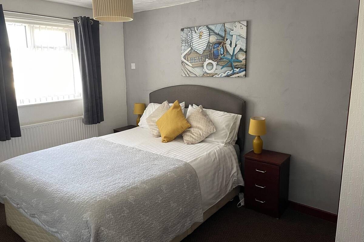 Un lit bien fait, avec une couette blanche et plusieurs oreillers, une table de chevet avec une lampe, et une œuvre d’art accrochée au mur représentant des éléments marins.