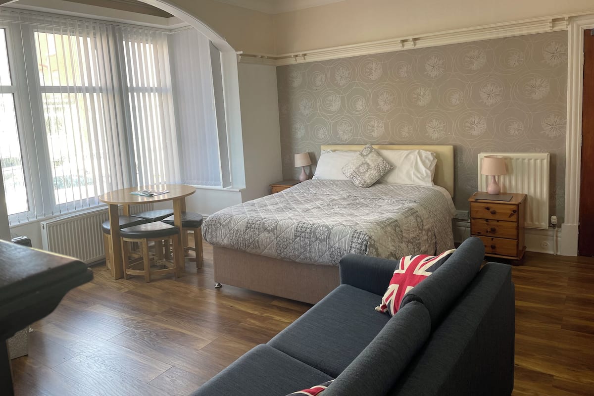Une chambre à coucher comprenant un lit, une table de chevet, une lampe et une vue sur l’extérieur par une fenêtre munie de stores.