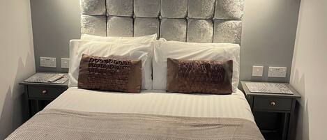 Habitación de hotel con un cabecero capitonado, dos mesitas de noche con lámparas, una cama con una colcha texturizada y dos almohadones estampados.
