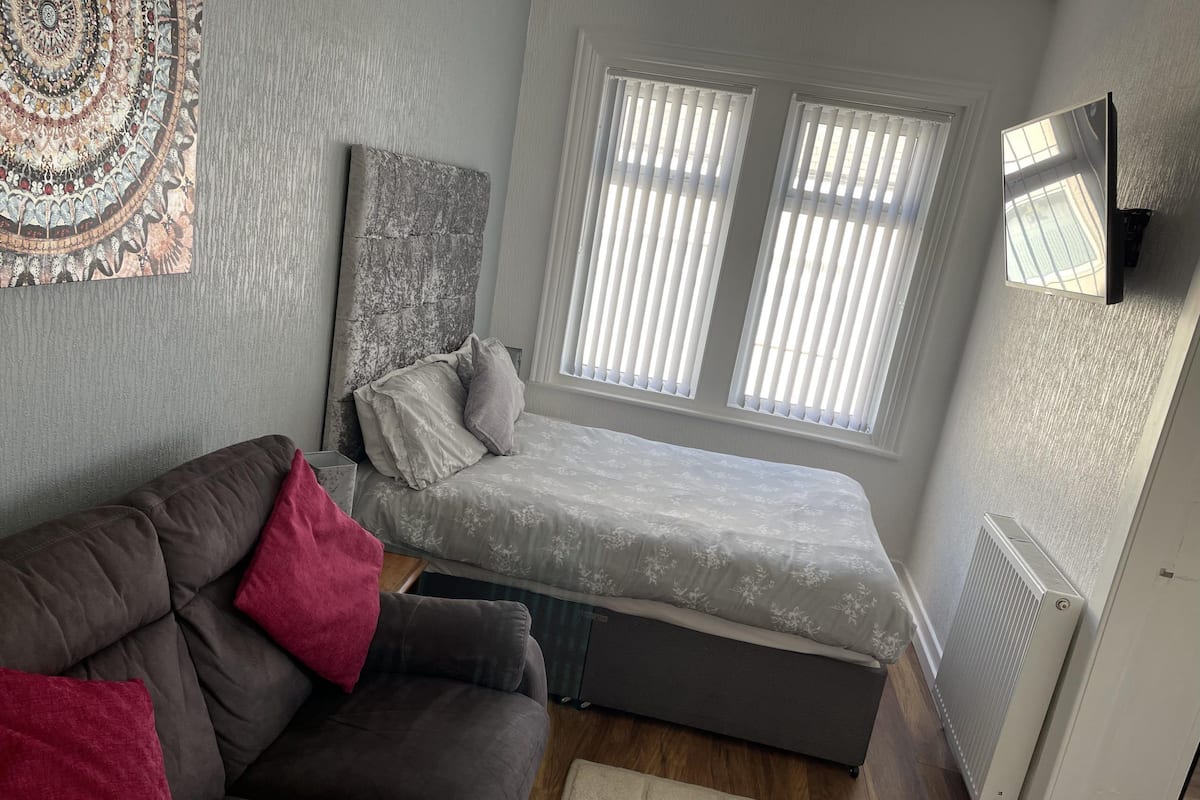 Une petite chambre à coucher comprenant un lit, un canapé, un radiateur et une fenêtre avec des stores.