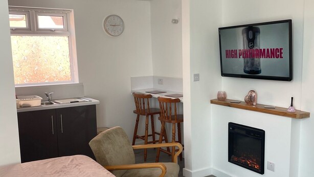 A compact living space with a kitchenette, a dining area with wooden chairs, a television mounted above a fireplace, and two beds with patterned bedding. - San Remo Apartments Blackpool (Blackpool)