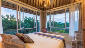 A bedroom with a large bed, multiple windows, and a view of greenery. - BoHo Bingin Beach (Pecatu)