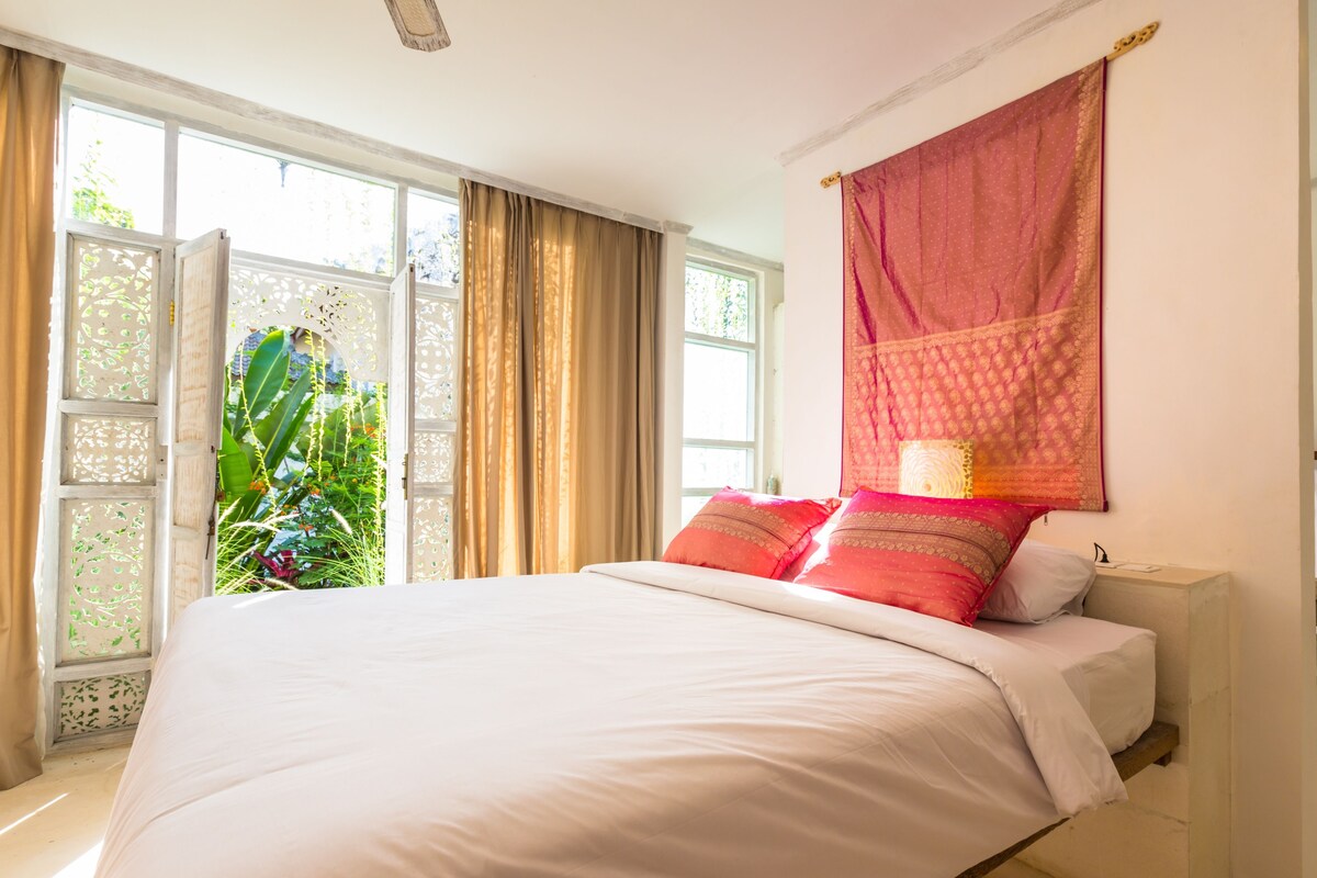 A bedroom with a large bed, a window with curtains, and a view of greenery outside.