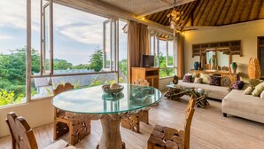 A spacious living area with a glass-top dining table, wooden furniture, and a sofa with cushions. Large windows offer a view of greenery outside. - BoHo Bingin Beach (Pecatu)